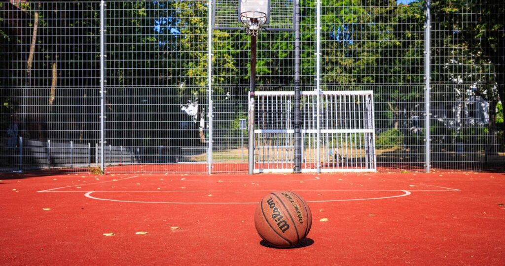 Leerer Outdoor-Basketballplatz mit rotem Belag, in der Mitte liegt ein brauner Wilson-Basketball, im Hintergrund Metallzaun und Basketballkorb vor Bäumen.